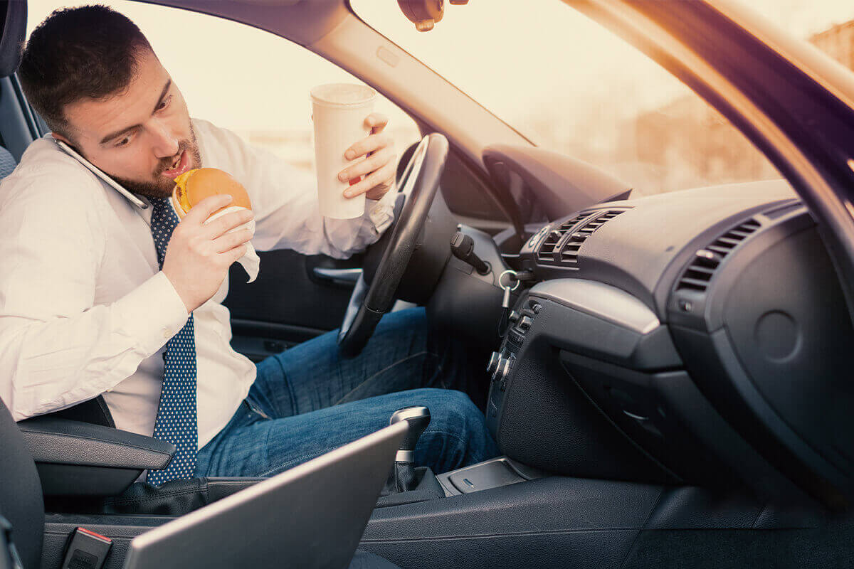 Man eating a hamburger, holding a drink, and talking on the phone while in the driver's seat